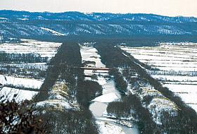 中央に1本の端が掛かった川が流れ、周りは雪で真っ白になっている平原と奥に山が見えている写真