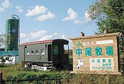 「中尾牧場」と書かれた木製の看板が手前にあり、奥に電車の車両が1両とサイロが見えている写真