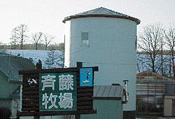 「斉藤牧場」と書かれた看板があり、奥に白い大きなサイロが見えている写真